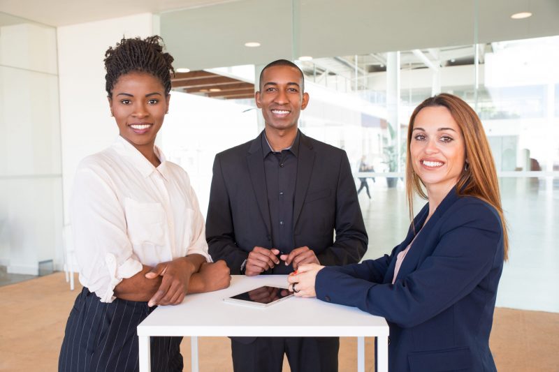 Cheerful successful team of three posing in office hallway. Business people in formal suits standing at counter and smiling at camera. Teamwork concept